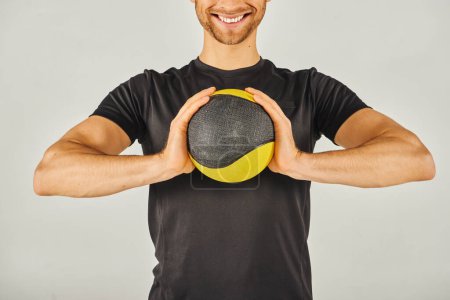 Jeune sportif en tenue active tenant énergiquement une boule jaune et noire vibrante dans un studio au fond gris.
