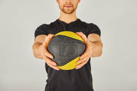 A young sportsman in active wear holds a yellow and black ball in a studio with a grey background.