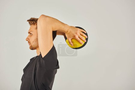 Jeune sportif en tenue active s'exerce vigoureusement avec une balle jaune dans un studio avec un fond gris.