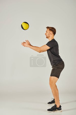 Un jeune sportif en tenue active attrape une balle jaune et noire dans un studio dynamique avec un fond gris.