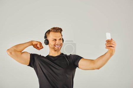 Foto de Un joven deportista en ropa activa tomando una selfie con su teléfono en un estudio con un fondo gris. - Imagen libre de derechos
