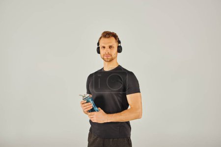Joven deportista en ropa activa, con auriculares, sostiene una botella de agua en un estudio con un fondo gris.