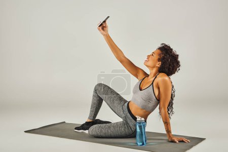 Eine lockige afroamerikanische Sportlerin macht ein Selfie, während sie in einem Studio mit grauem Hintergrund auf einer Yogamatte sitzt.