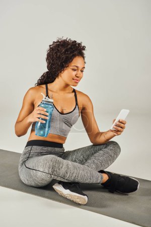 Une sportive afro-américaine frisée est assise sur un tapis de yoga, tenant une bouteille d'eau dans un studio de yoga avec un fond gris.