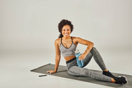 Lockige afroamerikanische Sportlerin in aktiver Kleidung sitzt auf einer Yogamatte, hält eine Wasserflasche in der Hand, mit grauem Studiohintergrund.