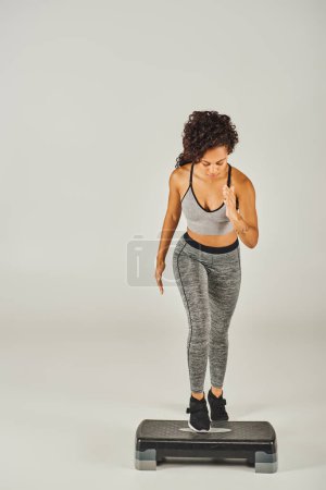 Curly Afro-American sportswoman in active wear stepping up on a white stepper in a studio.