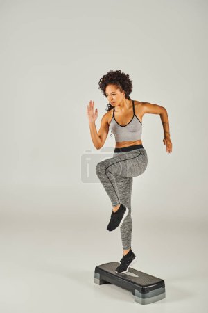 Foto de La deportista afro-americana rizada en ropa deportiva se ejercita graciosamente en un paso en un estudio con un fondo gris. - Imagen libre de derechos