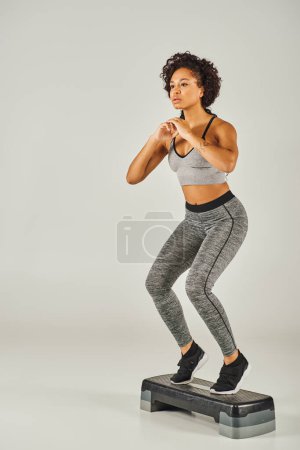 Lockige afroamerikanische Sportlerin in aktiver Kleidung tritt energisch auf einen Stepper in einem Studio mit grauem Hintergrund.