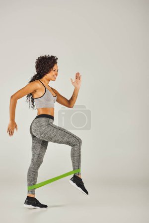 Photo for Curly African American sportswoman in active wear working out in studio with a resistance band against a grey background. - Royalty Free Image