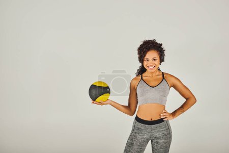 Curly African American sportswoman in active wear confidently holds a yellow and black ball in a studio setting.