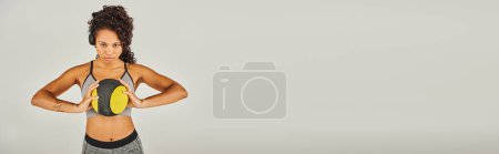 Téléchargez les photos : Femme sportive afro-américaine frisée en tenue active tenant une balle jaune vif dans ses mains dans un studio avec un fond gris. - en image libre de droit