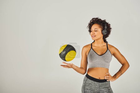 Téléchargez les photos : La sportive afro-américaine frisée en tenue active montre force et équilibre alors qu'elle tient une boule jaune et noire vibrante. - en image libre de droit
