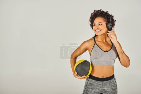 Lockige Afroamerikanerin in Sportbekleidung, trägt Kopfhörer und hält einen Ball,