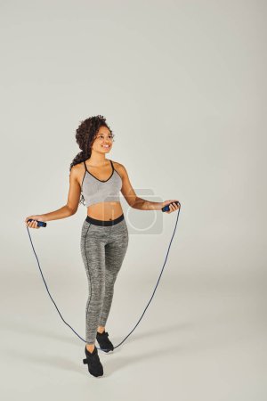 Curly African American sportswoman in active wear jumps with a skipping rope on a white background.