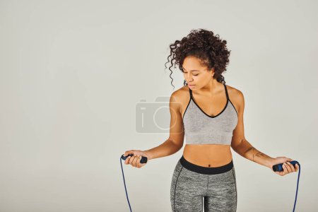 Foto de Una deportista afroamericana rizada en ropa activa sosteniendo una cuerda saltando frente a un fondo gris. - Imagen libre de derechos