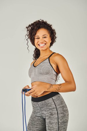 Foto de Una deportista afroamericana rizada en ropa deportiva sonríe mientras sostiene una cuerda saltando en un estudio sobre un fondo gris. - Imagen libre de derechos