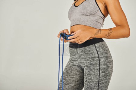 Foto de La deportista afroamericana en ropa deportiva sostiene enérgicamente una cuerda de salto en un estudio con un fondo gris. - Imagen libre de derechos