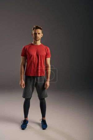 Photo for A man in a red t-shirt and grey shorts confidently strikes a pose in front of a serene grey background. - Royalty Free Image