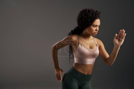 Femme de sport afro-américaine frisée en soutien-gorge de sport et leggings frappant une pose dans une séance photo avec une toile de fond grise.