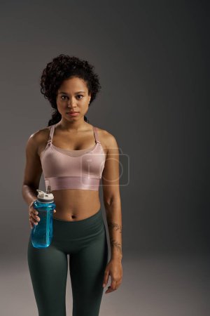 Eine lockige afroamerikanische Sportlerin in Tank-Top und Leggings, die während einer Trainingseinheit mit einer Wasserflasche feuchtigkeitsspendend wirkt.