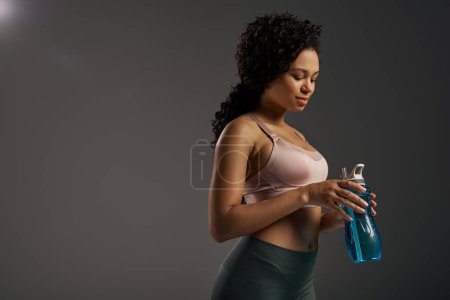 Eine lockige afroamerikanische Sportlerin in einem Sport-BH mit einer Wasserflasche, bereit für ein Training.