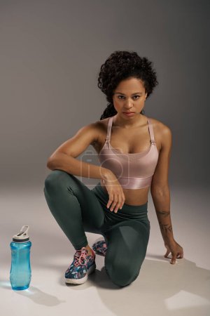 A young African American sportswoman in active wear gracefully crouching next to a vibrant blue water bottle in a studio.