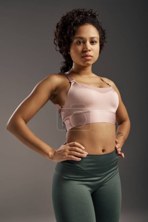 Téléchargez les photos : Femme de sport afro-américaine bouclée en soutien-gorge de sport rose et leggings verts travaillant en studio avec un fond gris. - en image libre de droit