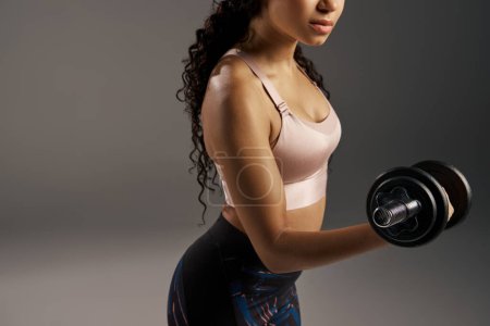 Photo for A curly-haired African American sportswoman in activewear works out with dumbbells in a grey studio background. - Royalty Free Image