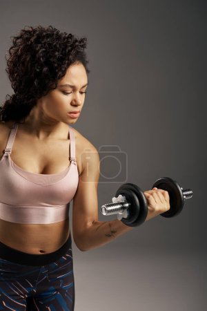 Photo for Curly African American sportswoman in active wear lifts a pair of dumbbells in a studio with a grey background. - Royalty Free Image