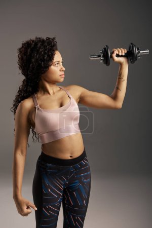 Photo for Curly African American sportswoman in active wear holding a dumbbell in front of a gray studio background. - Royalty Free Image