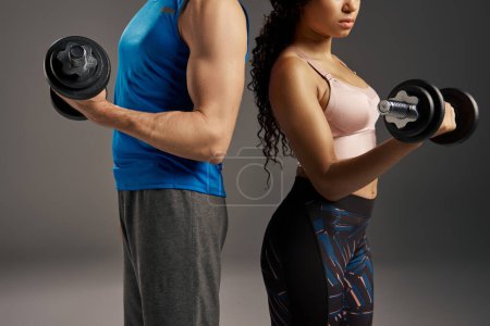 Young, fit multicultural couple in active wear holding dumbbells in front of a gray background.
