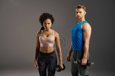 Multicultural fit couple in activewear strike poses with dumbbells against grey studio background.
