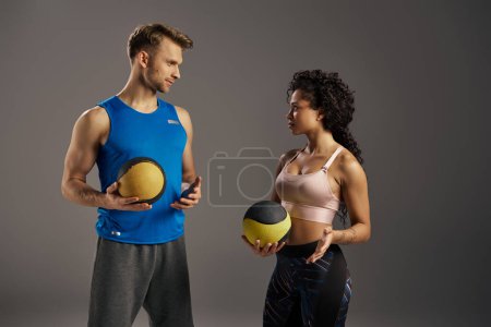 Young multicultural and fit couple in active wear standing next to each other, holding exercise balls in a studio setting.
