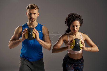 Young, fit man and woman in active wear lift balls together against a gray backdrop in a studio setting.