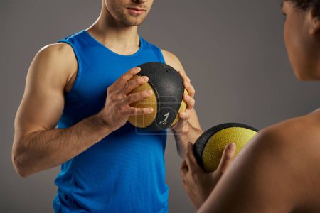 Multicultural, fit couple in active wear holding a yellow and black ball in a studio setting against a grey background.