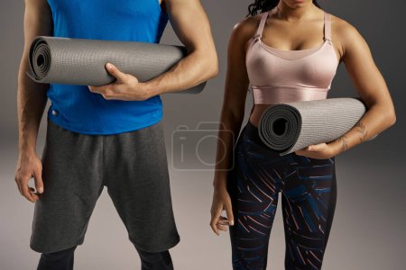 A young multicultural and fit couple in active wear holding a yoga mat in a studio setting against a grey background.