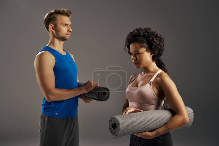 Multicultural couple in activewear stand next to a yoga mat, ready for a workout session in a studio on a grey background.