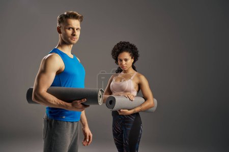 Photo for A young multicultural and fit couple in active wear holding a yoga mat, ready for their workout in a studio setting. - Royalty Free Image