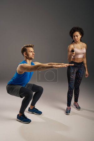 Photo for A young, multicultural couple in activewear perform squats on a grey background in a studio setting. - Royalty Free Image