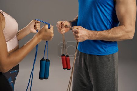 Young multicultural couple in workout gear, holding jump ropes, ready to start their fitness routine on a grey studio background.