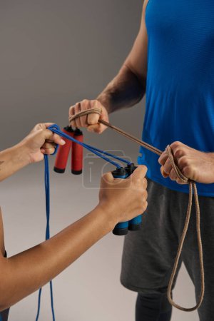 Téléchargez les photos : A man and a woman, both young and multicultural, in active wear, hold jump ropes in a fitness studio on a grey background. - en image libre de droit