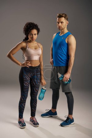 A young multicultural couple in active wear striking a powerful pose against a grey backdrop in a studio setting.