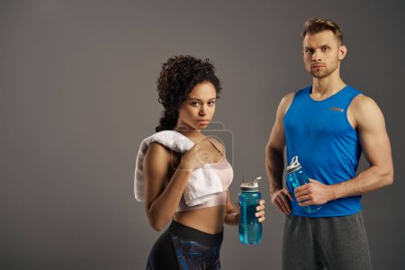 Foto de Una pareja multicultural y en forma en ropa activa posando con estilo con botellas de agua sobre un fondo gris de estudio. - Imagen libre de derechos