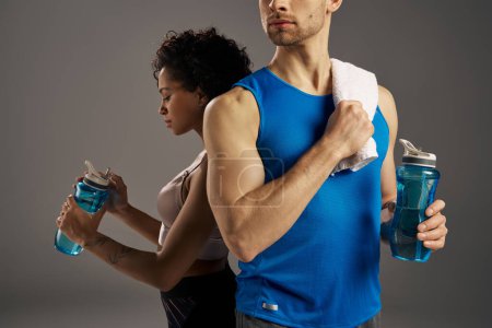 Multicultural couple in active wear holding water bottles in front of a gray background.
