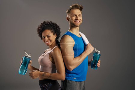 Ein multikultureller, fitter Mann und eine Frau in aktiver Kleidung posieren mit Wasserflaschen und zeigen gesunde Hydratationsgewohnheiten.