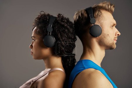 Photo for Young, multicultural couple in activewear, immersed in music, wearing headphones while working out in a studio against a grey backdrop. - Royalty Free Image