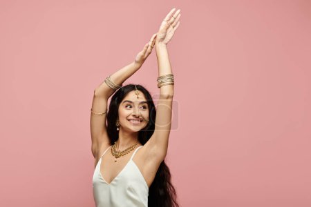 A graceful young indian woman poses in a white dress against a vibrant pink backdrop.