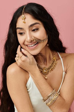 Téléchargez les photos : Jeune femme indienne aux cheveux longs et bijoux en or pose gracieusement sur un fond rose vif. - en image libre de droit
