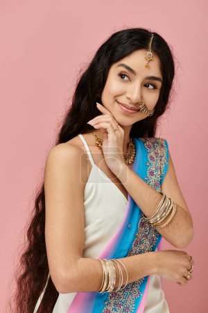 Young indian woman in blue and white sari poses gracefully on pink background.