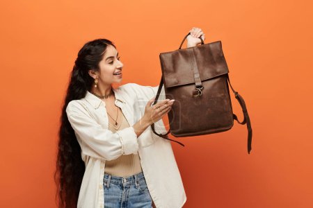 Beautiful indian woman holding a brown leather backpack against a vibrant orange background.
